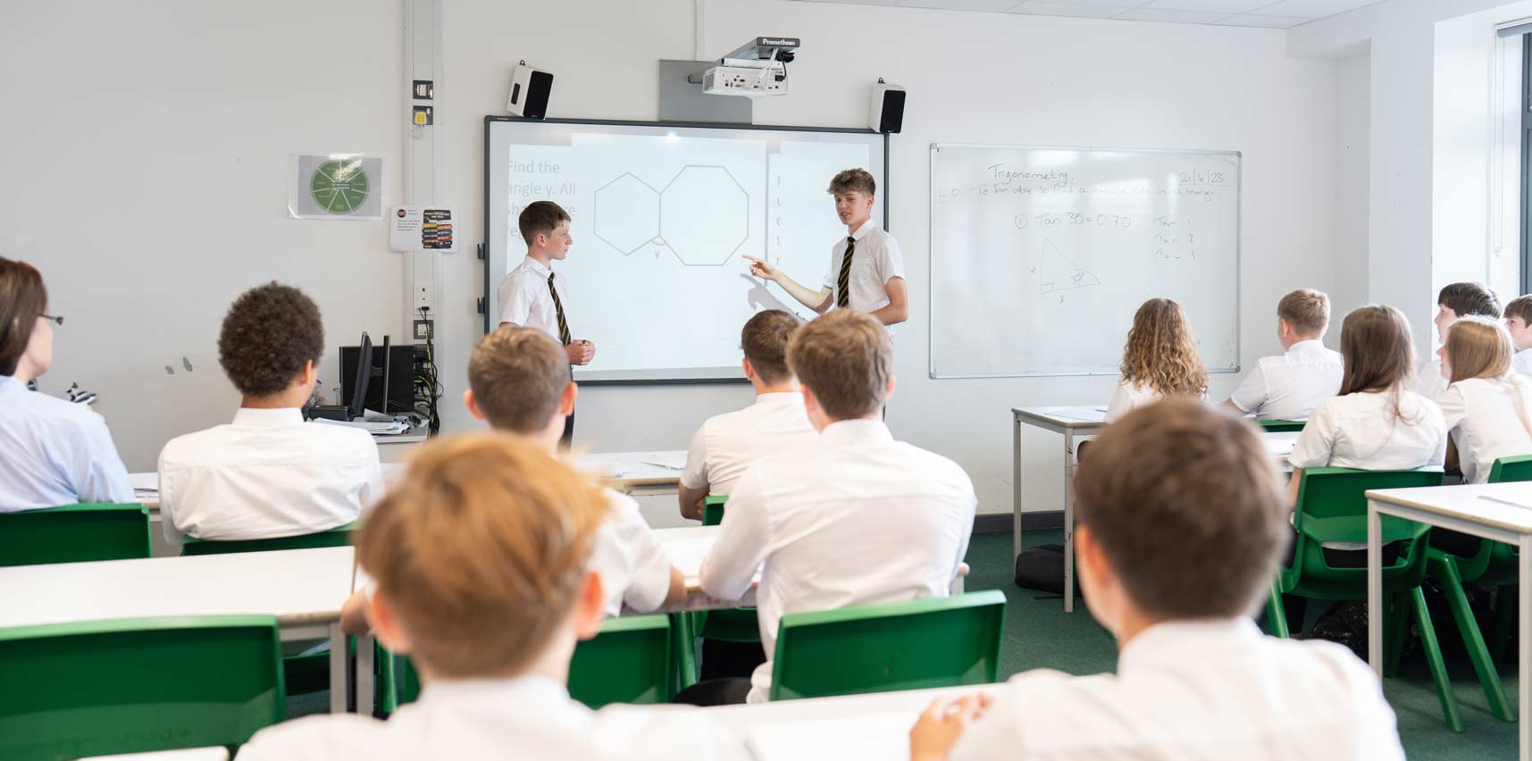 2 students stand at the front of the classroom and point at an image on the projector screen