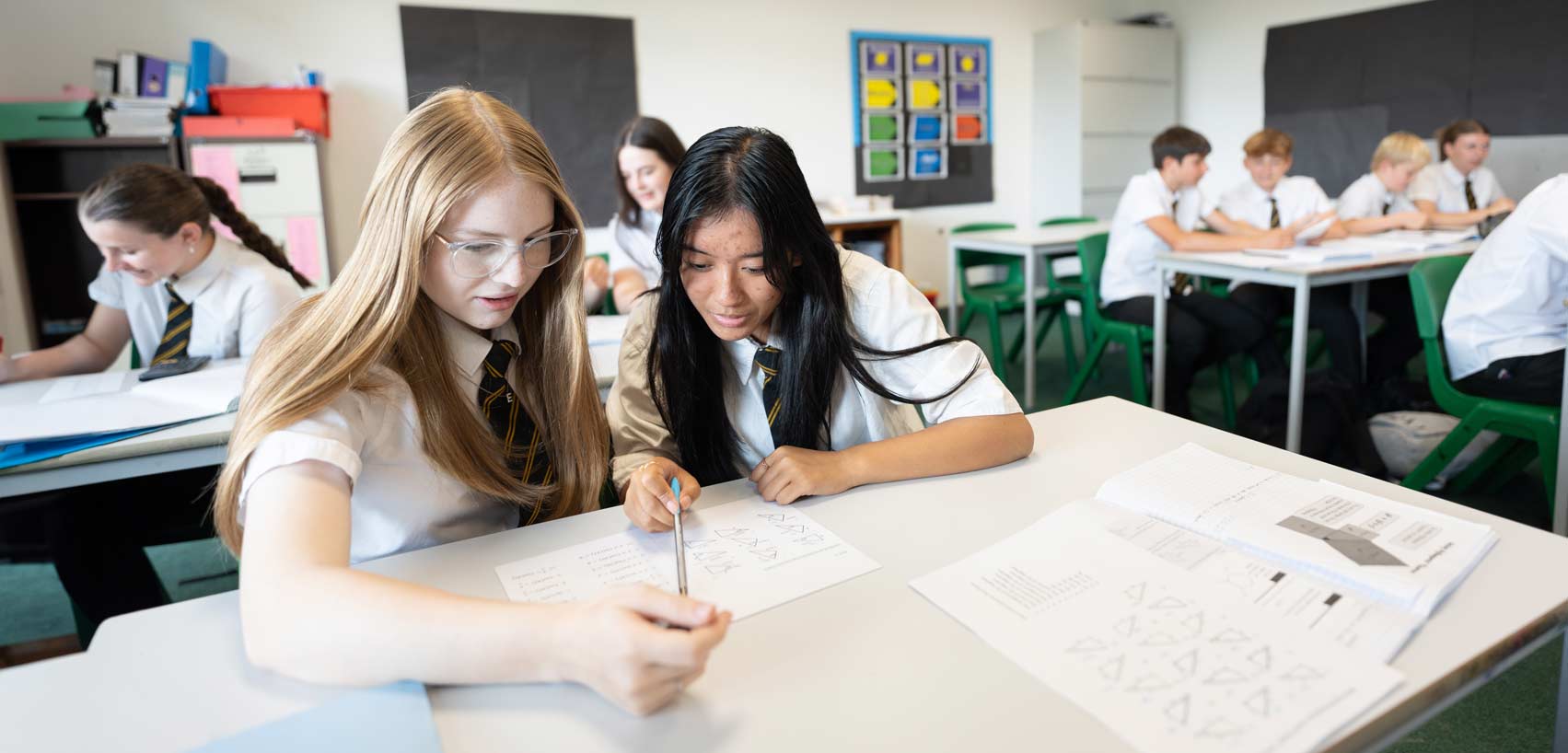 2 students sitting at a desk in a classroom. Both looking at a piece of paper on the desk.